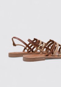Sandalias de cuero marrón con acentos en beige trenzado, correa ajustable, suela plana y suela de goma marrón claro, que muestran un diseño entrecruzado.
