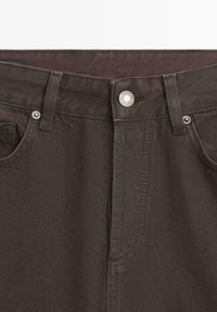 Tissu en denim marron avec une finition texturée, doté d'une fermeture à boutons sur le devant et de poches latérales discrètes. Les accents comprennent des éléments métalliques.