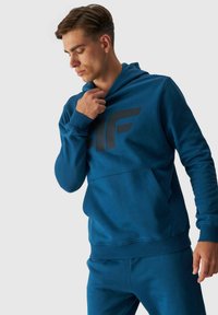 Sudadera azul de tejido suave, con un gran logo negro en la parte frontal, bolsillo tipo canguro y capucha ajustable con cordón.