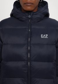 Mornarice pufast jakna z zadrgo in kapuco. Ima prešite panele, mehak tekstil in detajle z logotipom na prsih.