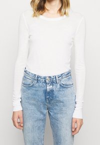Weißes Langarmshirt mit Rundhalsausschnitt, kombiniert mit hoch taillierten, hellblauen Jeans aus Denim. Das Shirt hat schmale Ärmel und eine weiche Textur.