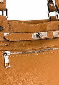 Détail d'un sac à main en cuir texturé beige avec des finitions en métal argenté, fermeture éclair et coutures contrastées le long des bords.