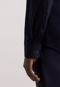 Camicia blu navy con bottoni, caratterizzata da una texture liscia, polsini regolabili con bottoni blu e un design su misura.