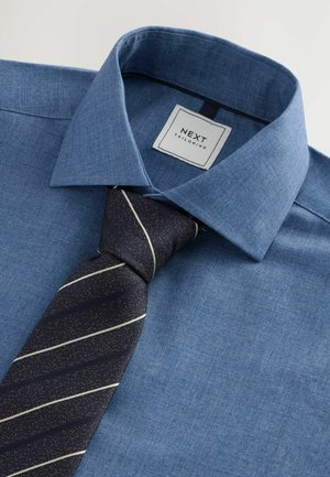 Jasnoniebieska koszula z guzikami wykonana z teksturowanej tkaniny, z ciemnogranatowym krawatem w metaliczne srebrne paski. Metka mówi "NEXT TAILORING."