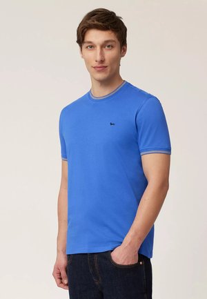 Giovane uomo che indossa una T-shirt a maniche corte di un azzurro brillante con un piccolo logo nero di un cane, jeans scuri, in piedi davanti a uno sfondo chiaro e neutro.