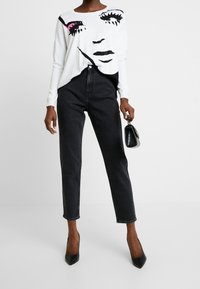 Bílá košile s dlouhým rukávem zdobená grafickým motivem obličeje v černé a růžové barvě, kombinovaná s černými džíny s vysokým pasem a rovnými nohavicemi a špičatými podpatky.