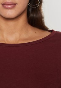 Burgundi-paita, jossa on leveä, pyöreä kaula-aukko. Sujuva rakenne ja istuva leikkaus. Ominaispiirteenä hienovarainen kiilto ja lyhyet hihat.