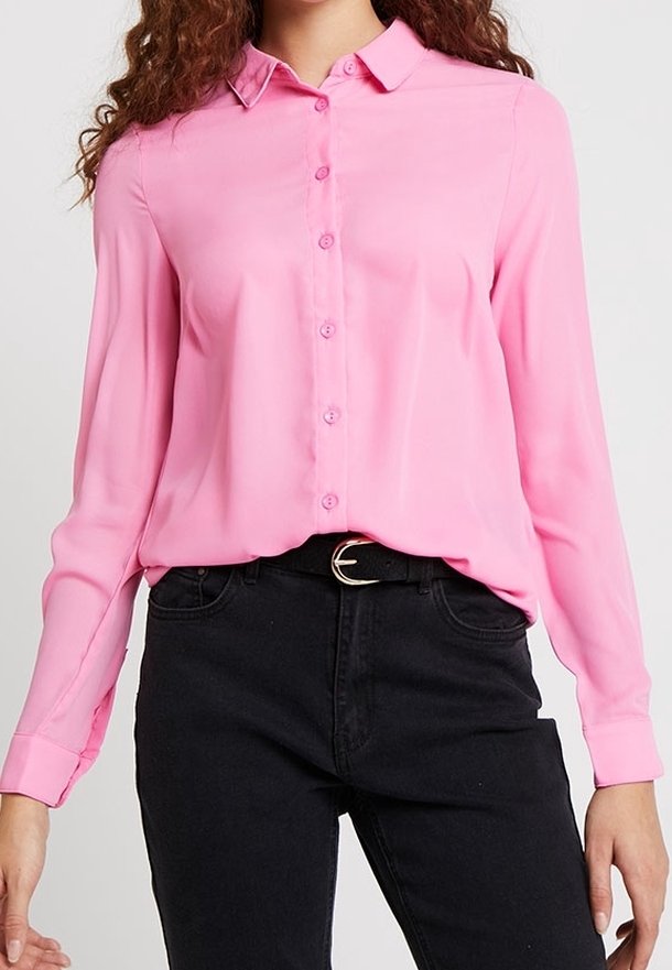 Femme portant une chemise rose à manches longues boutonnée, rentrée dans un jean noir taille haute avec une ceinture noire.