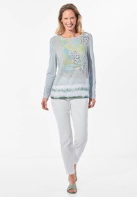 Langärmliges Oberteil mit einer Mischung aus Pastellmustern, Streifen und einer glatten Textur, kombiniert mit weißen enganliegenden Hosen und grünen Sandalen.