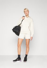 Kremhette med leopardmønsterdetaljer, matchende kremshorts, svart sportstasske, svarte joggesko med lilla detaljer og svarte sokker.