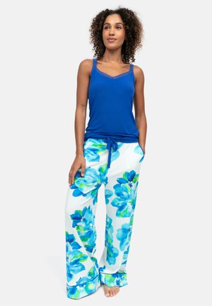 Frau steht barfuß, trägt ein blaues ärmelloses Oberteil und weiße, weit geschnittene Hosen mit großem blauem Blumenmuster und Tunnelzug an der Taille.