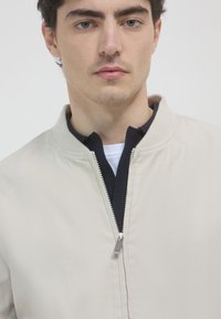 Giovane uomo con capelli ricci corti, che indossa una giacca beige con zip sopra un colletto nero e una camicia bianca, su uno sfondo neutro.