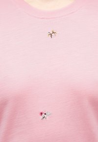 Ανοιχτό ροζ ύφασμα με μικρά κεντητά λουλουδένια σχέδια κοντά στη γραμμή του λαιμού και στο κέντρο του στήθους.