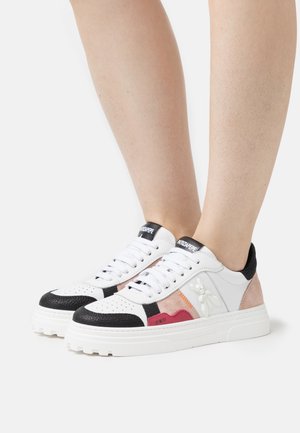 Hvide lavtop-sneakers med sorte, pink og røde accenter, båret på bare fødder mod en hvid baggrund.