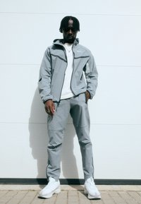 Γκρι φούτερ με φερμουάρ και κουκούλα, ταιριασμένα γκρι παντελόνια. Και τα δύο διαθέτουν λεία υφή, με αντίθετους σκούρους τόνους. Λευκά αθλητικά παπούτσια.