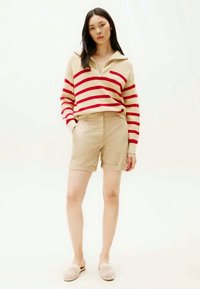 Young woman wearing beige shorts, beige and red striped long-sleeve pullover, and beige slip-on woven shoes, standing with hands in pockets.