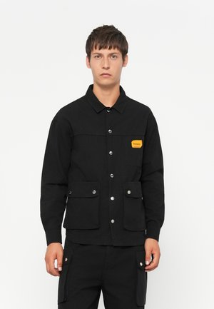 Black jacket with a button-up front, two front pockets with flaps, and a small orange label on the left chest. Flat collar, cotton material.
