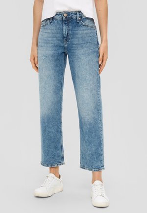 Femme portant un jean droit taille haute bleu clair avec des effets légèrement usés et des baskets blanches, debout devant un fond neutre.