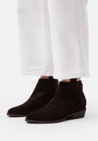 Stivaletti in suede marrone con punta affusolata, cuciture a contrasto e linguette. Indossati con pantaloni bianchi larghi.