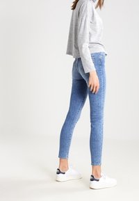 Jeans de denim azul ajustados con una cintura de altura media, un ligero desgaste y un diseño de cinco bolsillos, combinados con una camiseta de manga larga gris y zapatillas blancas.