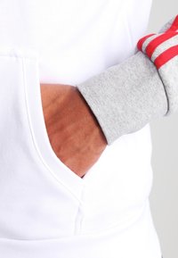 Sweatshirt blanc avec une poche avant, poignets de manches gris et rayures rouges aux épaules ; matériau en coton avec une texture lisse.