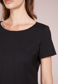 Robe noire à manches courtes avec un décolleté arrondi. Caractérisée par des coutures verticales et une texture lisse. Gros plan sur les détails de l'épaule et du décolleté.
