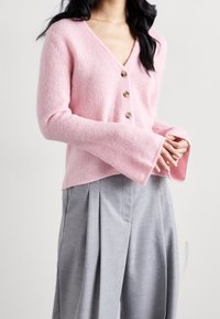 Rosa stickad kofta med V-ringning, fyra runda bruna knappar och trumpetärmar. Kombinerad med ljusgrå, veckade byxor.