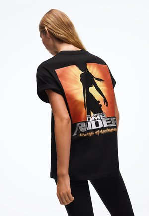 Pitkätukkainen nainen pukeutunut mustaan Tomb Raider -t-paitaan, jossa on siluettikuva ja oranssi tausta, nähtynä takaa.