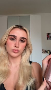 Jonge vrouw met lang blond haar houdt een roze metallic fles vast, staande binnen met witte kasten en een fotocollage op de achtergrond.