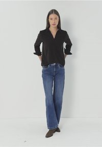 Chemise noire à col, manches longues retroussées ; jeans larges bleu avec un léger délavage ; bottines brunes à la cheville ; fond blanc uni.