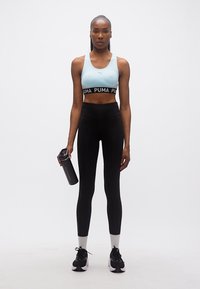 Αθλητική γυναίκα με ανοιχτό μπλε αθλητικό σουτιέν και μαύρο κολάν κρατώντας ένα μαύρο παγούρι νερού, στέκεται μπροστά σε απλό λευκό φόντο.