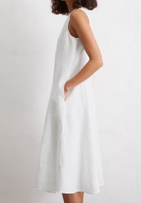 White linen dress with a sleeveless design. Features side pockets and a flared silhouette, reaching mid-calf with a smooth, textured finish.