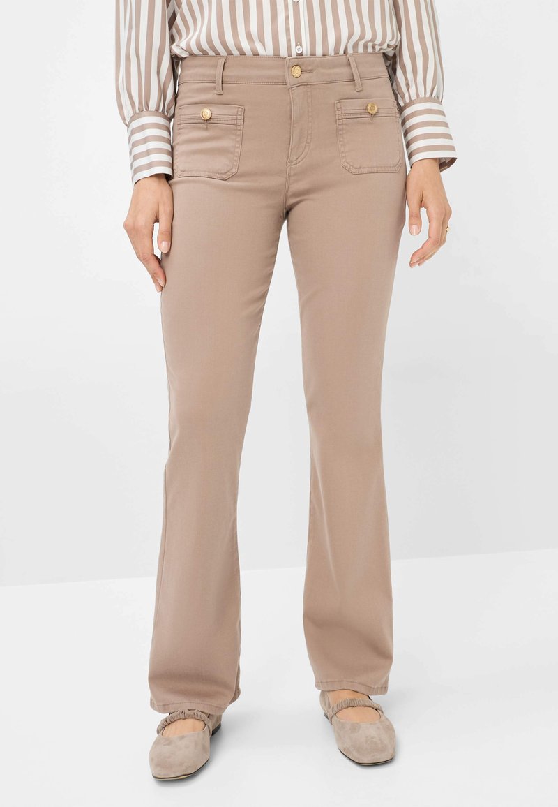 Beige Schlaghose mit Vorderaschen und goldenen Knopfakzenten. Der Stoff wirkt glatt mit einem subtilen Glanz; kombiniert mit hellen Schuhen.
