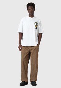Hvid bomulds T-shirt med en sort og guld pokal grafisk design på brystet, parret med brune fløjlsbukser med vidde og sorte sko.