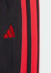 Czarne spodnie sportowe z czerwonymi paskami i haftowanym czerwonym logo. Tkanina jest miękka o teksturowanym wykończeniu.