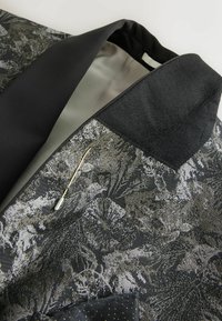 Textured black and silver patterned blazer with a satin lining, pointed lapel, and gold pin accent on the collar. Modern and structured design.