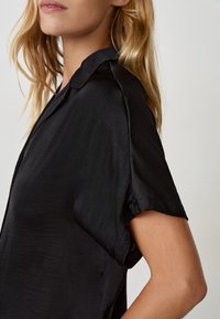 Maglietta nera a maniche corte realizzata in tessuto lucido, con colletto classico e vestibilità rilassata, caratterizzata da spalle pronunciate e texture morbida.