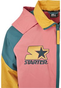 Veste à blocs de couleurs avec un corps rose, une manche bleu sarcelle, et un col jaune, arborant un logo Starter jaune et noir sur la poitrine.
