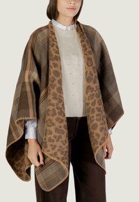 Kobieta nosząca brązowy szal w kratę i w leoparda na białym swetrze z perłowymi zdobieniami oraz ciemno-brązowych spodniach.
