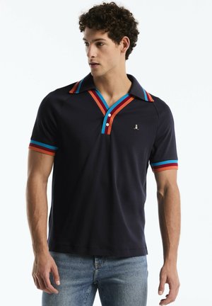 Young man wearing a navy polo shirt with red, blue, and orange stripes on collar and sleeves, paired with light blue jeans.