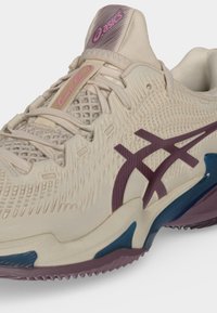 Μπεζ αθλητικό παπούτσι Asics με υφασμάτινη υφή, μωβ λογότυπο, ροζ κείμενο "Court FF" και μπλε και μωβ λεπτομέρειες στη σόλα.