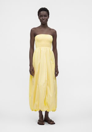 Femme portant une robe longue jaune sans bretelles avec des rayures verticales et des tongs marron, debout devant un fond uni.