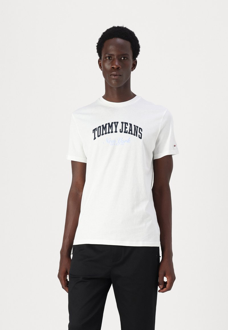 Fehér pamut póló, amelyen a "TOMMY JEANS" és a "New York" feliratok sötétkék színben szerepelnek. Rövid ujjú és kerek nyakú. Fekete nadrággal viselve.