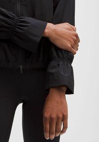 Sort letvægtsjakke med indsamlede manchetter, udstyret med lynlås i taljen og glat, blank tekstur. Laget over sorte leggings.