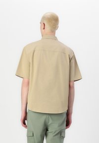Chemise à manches courtes en tissu beige clair, avec un design épuré et un ourlet arrondi. La vue de dos montre un col simple et une texture douce.