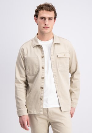 Jonge man met krullend haar, gekleed in een beige overhemdjas over een wit T-shirt en bijpassende beige broek, staand tegen een effen achtergrond.