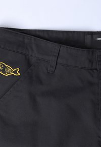 Zwarte stoffen broek met een gladde textuur, voorzien van een gele geborduurde vissenlogo en een label met de tekst "FORMER" op de tailleband.
