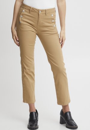 Pantalons en mélange de coton beige avec une coupe droite, dotés d'accents de boutons sur le côté et d'une texture lisse. Associés à des chaussures à talons noires.