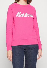 Sweatshirt rose avec lettrage "Barbour" blanc, fabriqué en tissu doux, il présente un col rond et des poignets côtelés, associé à un jean foncé.