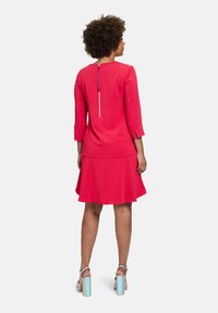Rotes, mittellanges Kleid mit Dreiviertelärmeln und einem Reißverschluss auf der Rückseite. Hat einen ausgestellten Rock und eine glatte, matte Textur. Silberne High Heels mit irisierenden Akzenten.